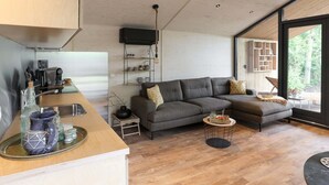 A modern living space with a kitchen area, a gray sectional sofa, a small round table, and a large window offering a view of greenery. - Cosy Holiday Home With Private Sauna (Wissenkerke)