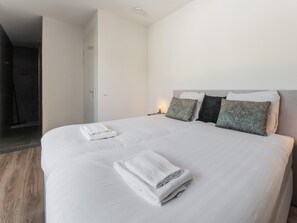 A neatly made bed with white linens and folded towels, a bedside table with a lamp, and a door leading to a bathroom. - Villa in Zeewolde for 6 Person (Zeewolde)