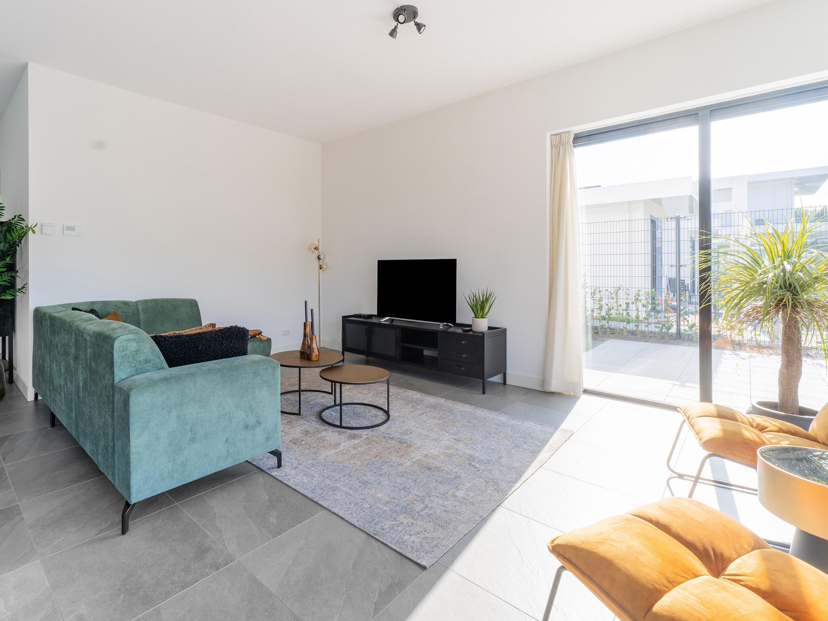 A modern living room with a teal sofa, a black entertainment unit, a flat-screen TV, a small round table with a vase, and a large sliding glass door leading to a balcony with a plant.