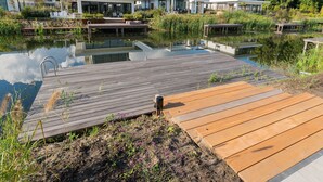 A modern residential area with a wooden dock extending into a calm water body, surrounded by lush greenery and contemporary houses. - Villa in Zeewolde for 6 Person (Zeewolde)