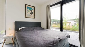 Une chambre moderne avec un grand lit, une table de chevet avec une lampe et une fenêtre donnant sur un paysage verdoyant et des bâtiments.