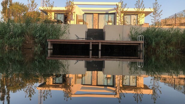 Une maison moderne avec une grande fontaine à l’avant, entourée de verdure et sous un ciel dégagé.