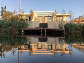 A modern house with a large water feature in front, surrounded by greenery and a clear sky.