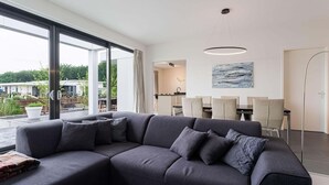 Un salon moderne avec un canapé d’angle gris, un coin repas comprenant une table et des chaises, et une vue sur l’extérieur.