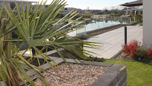 A modern outdoor area with a pool, palm plants, and a paved walkway. - Exclusive Villa With Boat Terrace (Zeewolde)