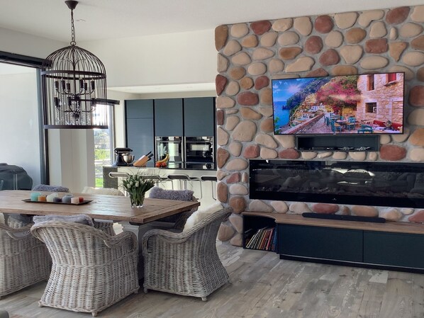 A modern living room with a stone wall, a flat-screen TV, wicker furniture, and a kitchen in the background. - Exclusive Villa With Boat Terrace (Zeewolde)