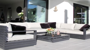 A modern outdoor seating area with a sofa and chairs, a coffee table with drinks, and a glass door leading to a room. - Exclusive Villa With Boat Terrace (Zeewolde)
