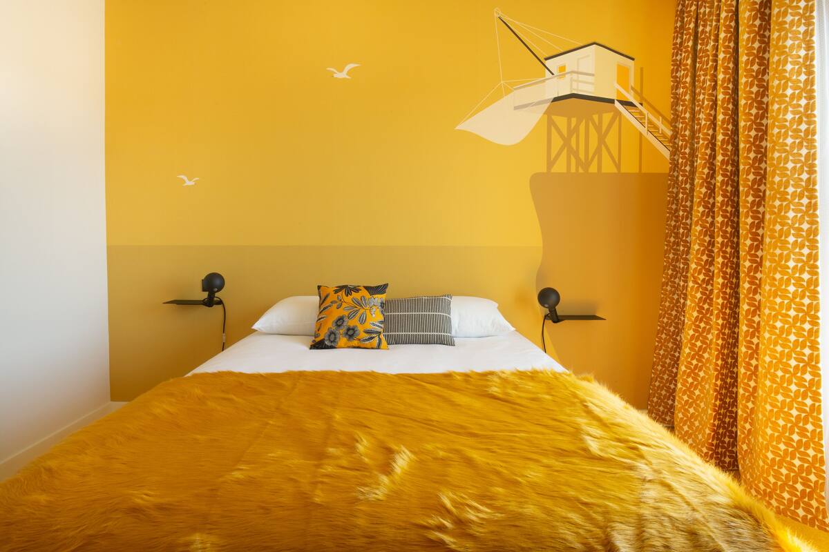 Cama com manta amarela, duas almofadas e um mural na parede.