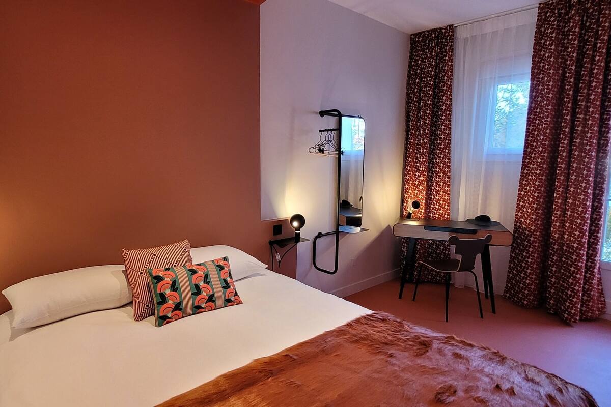 Quarto de hotel com cama, escrivaninha, cadeira, espelho e janela com cortinas.