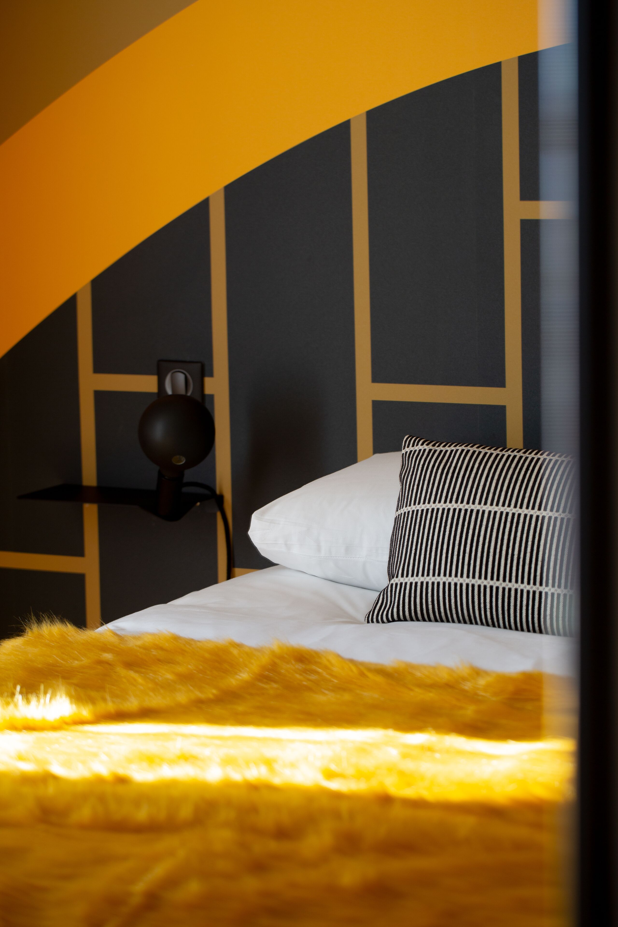 A bed with a white duvet and a striped pillow, a black lamp on a shelf, and a yellow furry blanket.