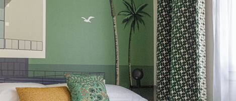 Ein Schlafzimmer mit grüner Wand, einem Bett mit grün-weißem Musterkissen, einer flauschigen, grünen Decke und einem Wandgemälde mit Palmen.