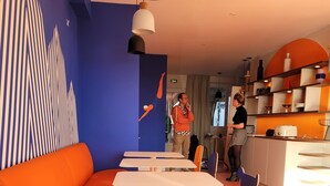 A modern interior with a blue and orange color scheme, featuring a dining area with white tables and chairs, a kitchen area with a toaster, and a seating area with orange sofas. - SEAKUB HOTEL (Royan)