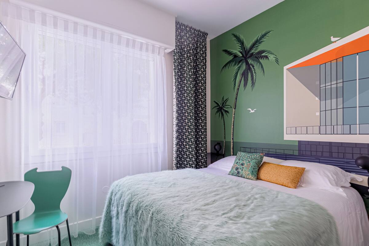 Quarto moderno com cama, uma cadeira, uma mesa e parede verde com um mural de palmeira.
