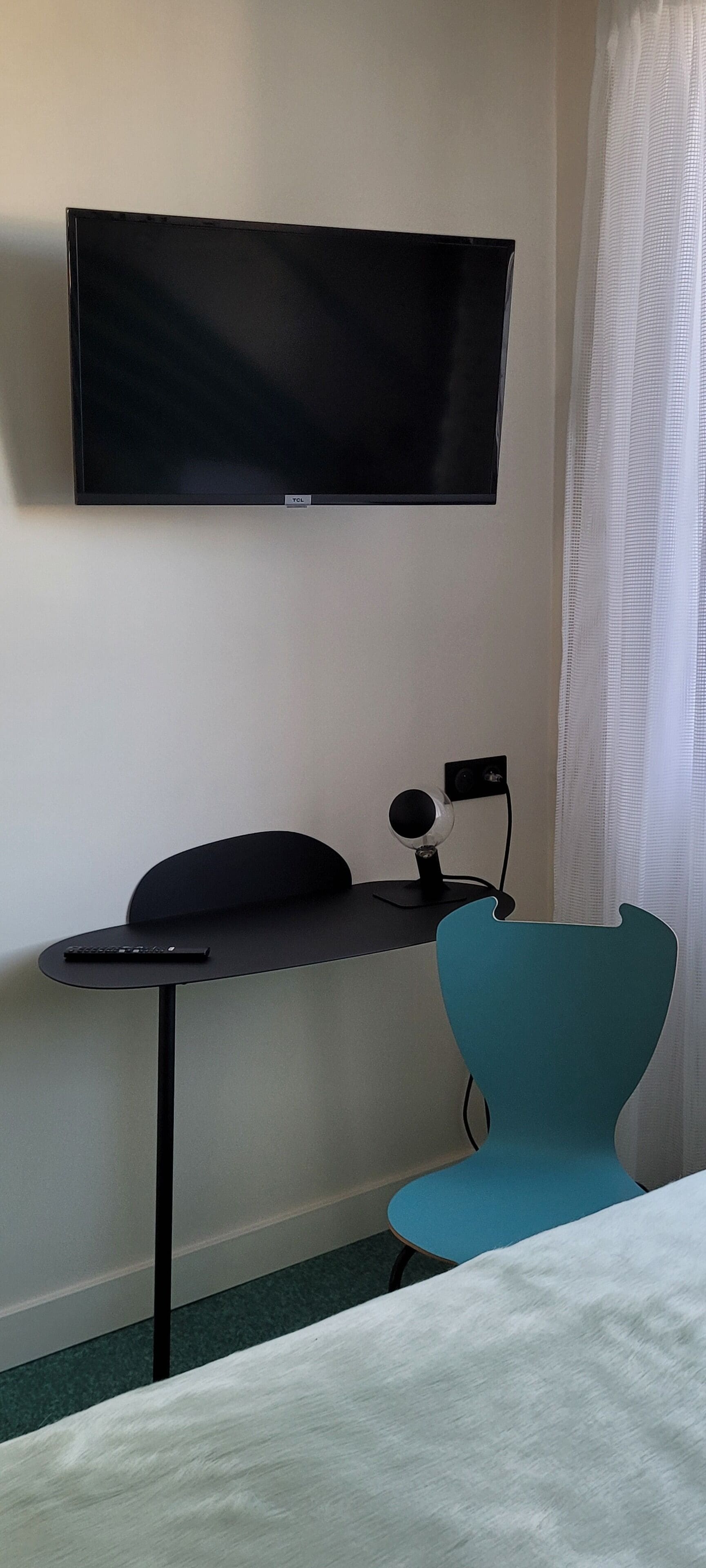 A modern room with a flat-screen TV mounted on the wall, a desk with a remote control, and a teal chair.