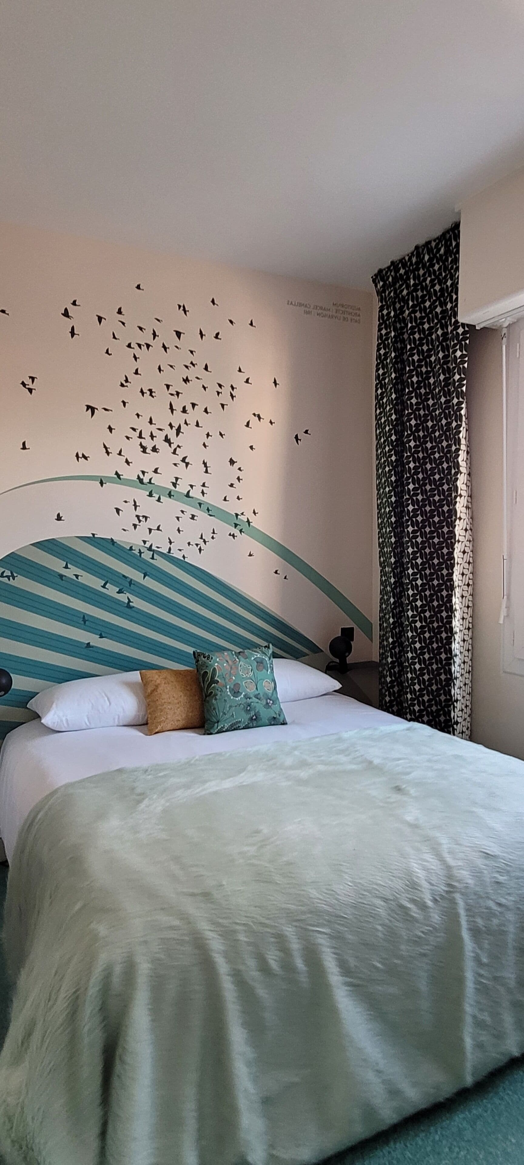 Quarto com cama, almofadas, parede com decalque de pássaro e cortina.