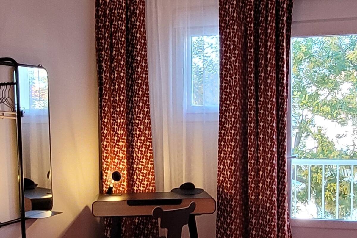 Quarto com cama, escrivaninha e cadeira. Há uma janela com cortinas.