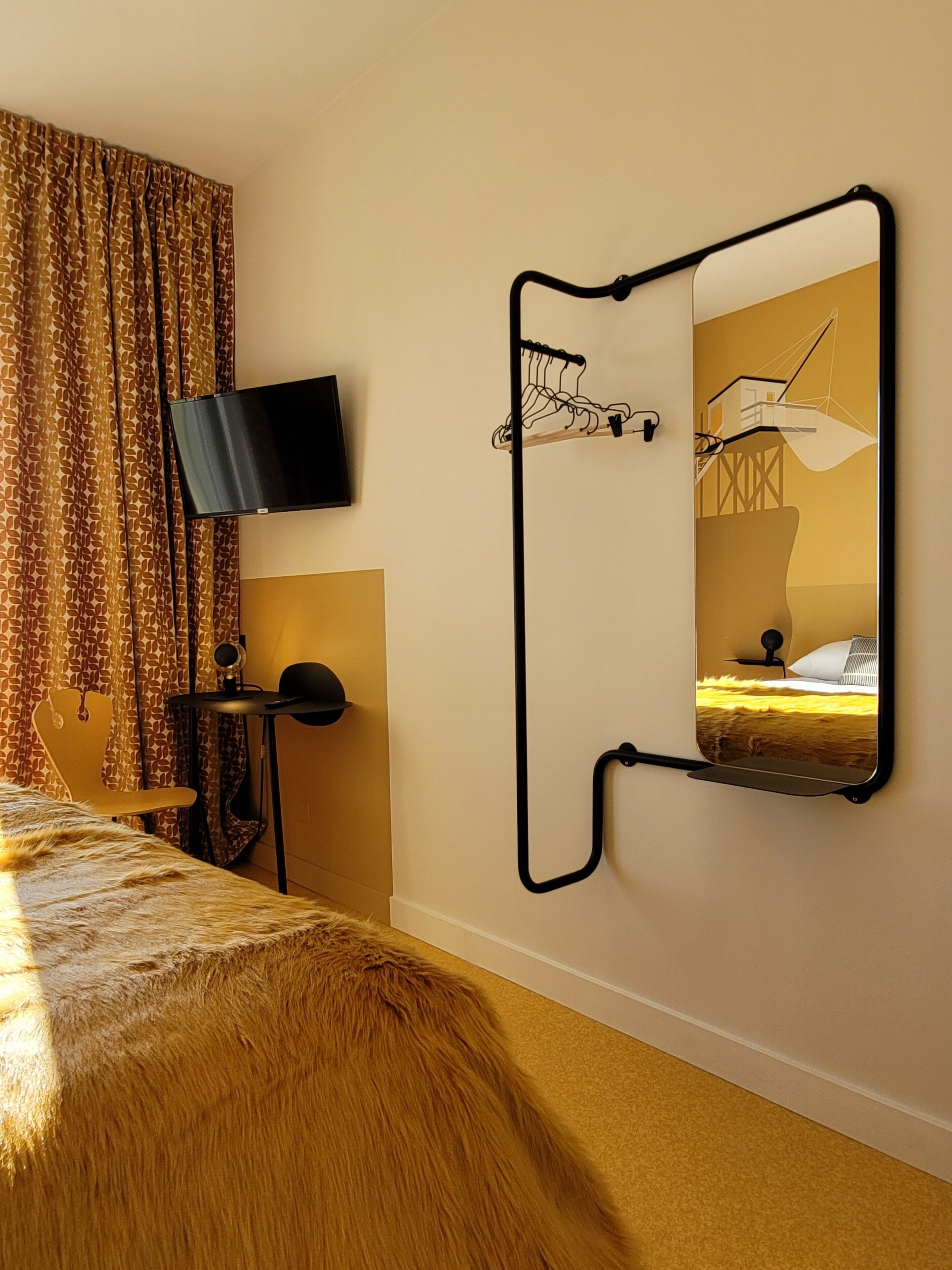 Een hotelkamer met een bed, een wandtelevisie, een spiegel met een bijzondere lijst en een raam met gordijnen.