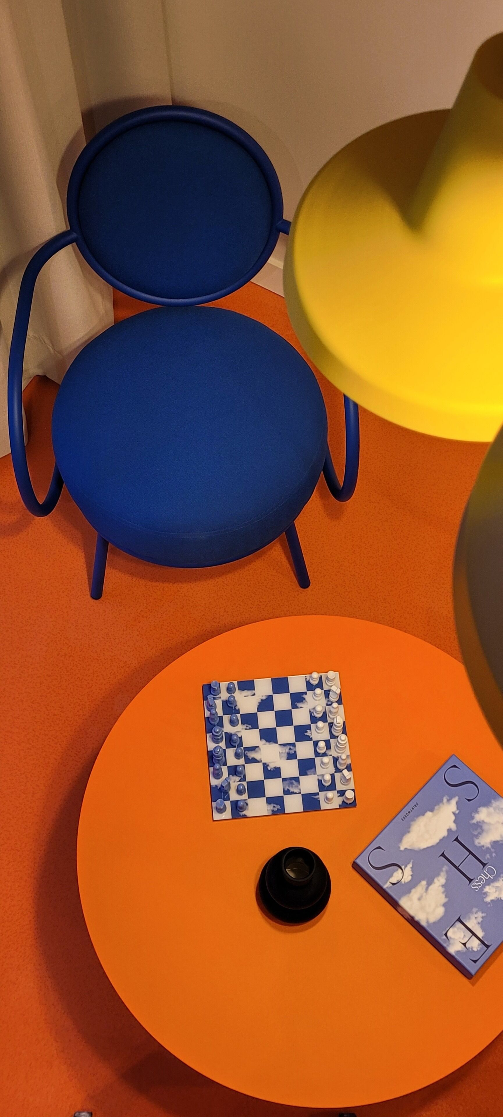 A room with two blue chairs, an orange table with a chessboard and a book, and a yellow lamp.