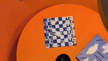 Une pièce avec deux chaises bleues, une table orange sur laquelle se trouvent un jeu d’échecs et un livre, et une lampe jaune.