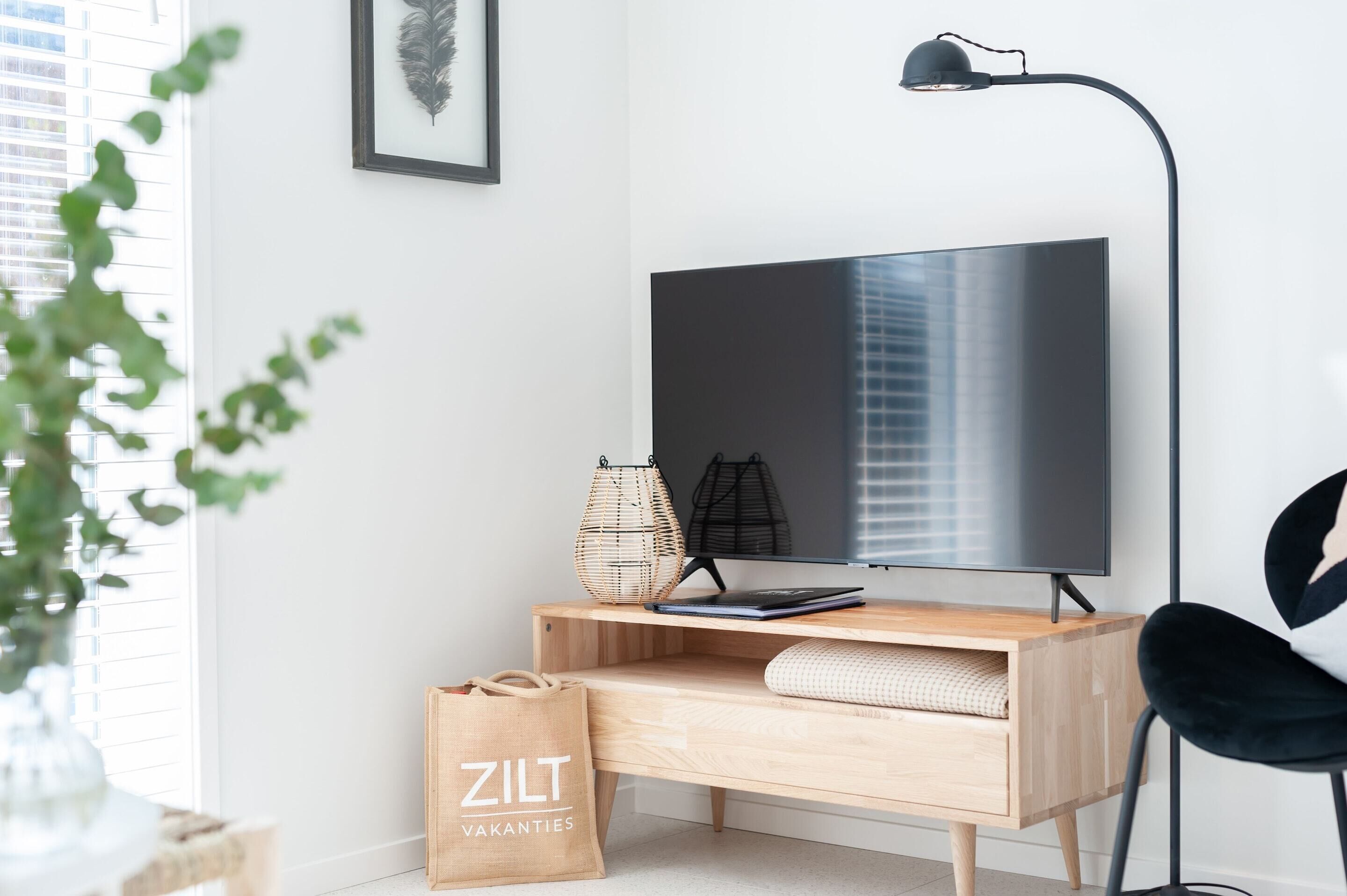 A modern living room with a flat-screen TV, a wooden desk, a black chair, a floor lamp, and a wicker basket.