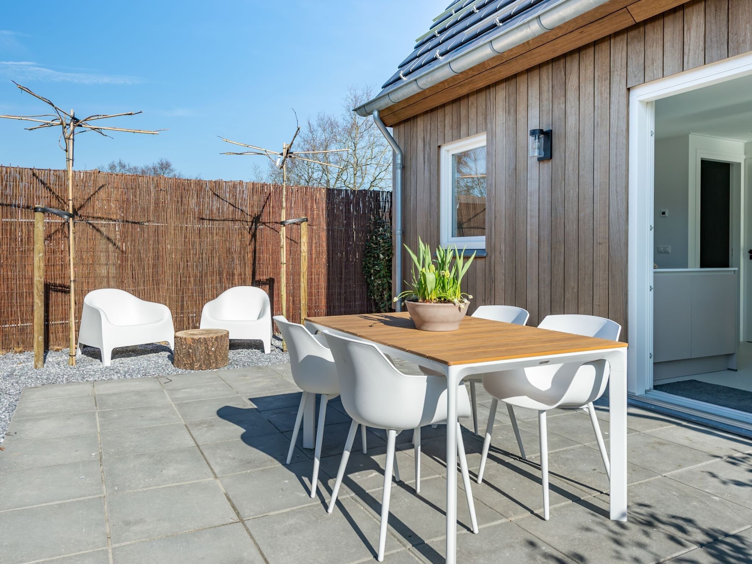An outdoor patio with a wooden table and white chairs, a potted plant, and a bamboo fence.