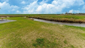 A grassy field with a small stream, a wooden bench, and a distant building. - Holiday Home in Zeeland Near Nature Reserve (Scherpenisse)