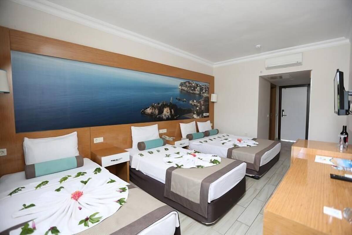 Habitación de hotel con tres camas, cabecera de madera, un cuadro grande con una escena costera, un escritorio y un espejo.