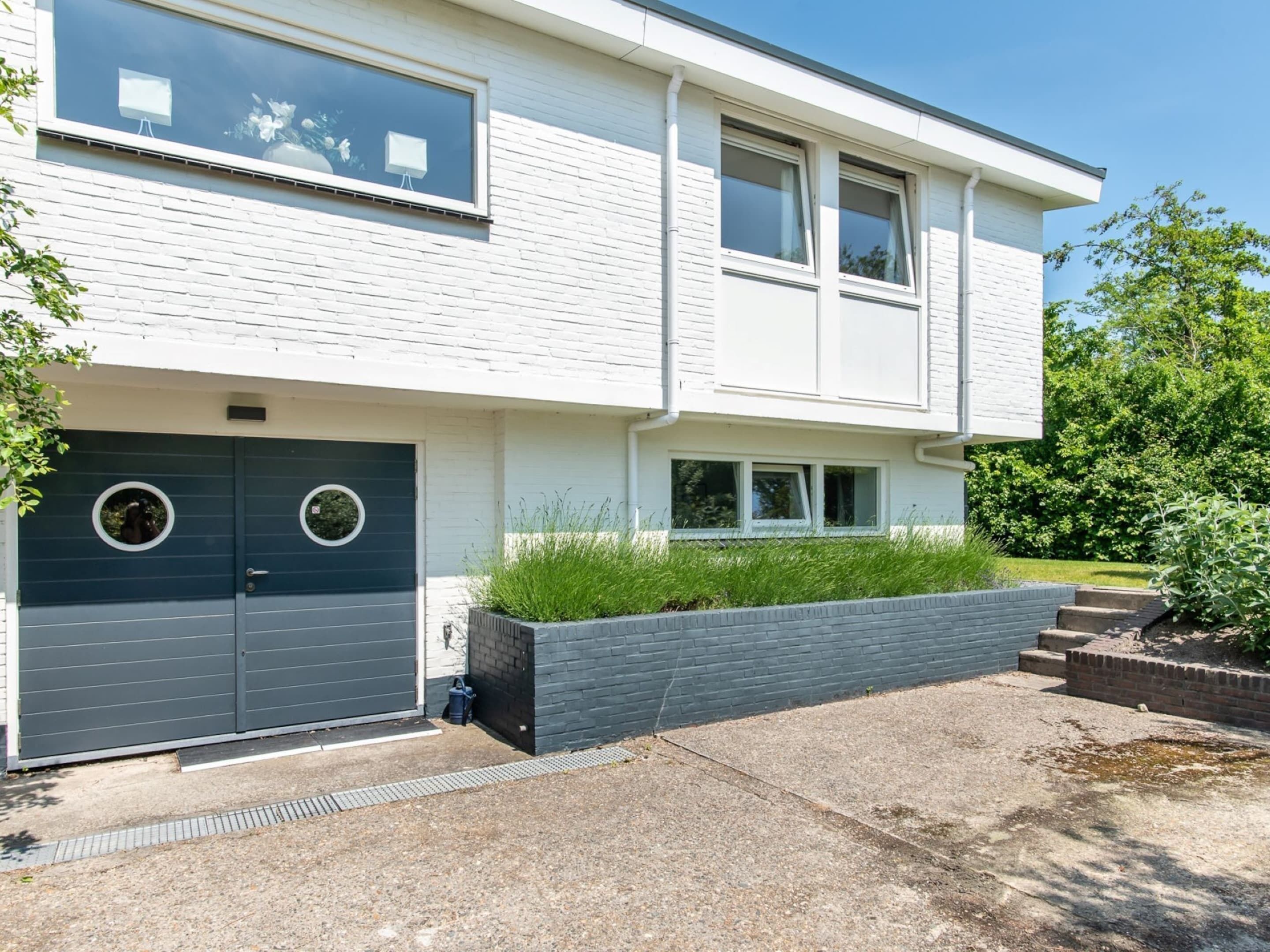 Een modern huis met twee verdiepingen, een donkerblauwe garageklep, een kleine voortuin met gras en een bakkenplanter, en een helderblauwe lucht.