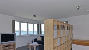 Ein modernes Wohnzimmer mit Fernseher, Sofa, Couchtisch, Bücherregal und Blick aufs Wasser.