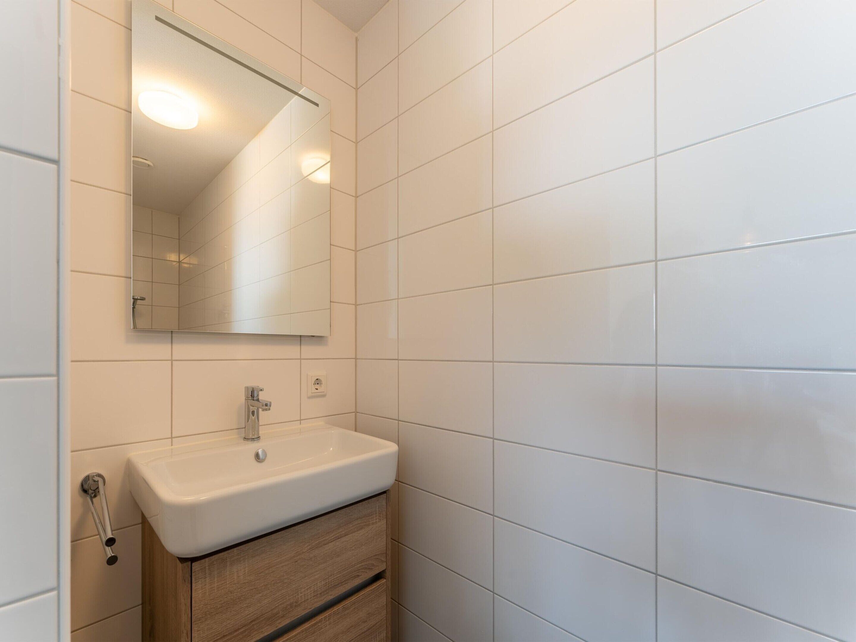 A bathroom with white tiles, a white sink, a mirror, and a wooden cabinet.