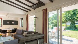 En moderne stue med en hjørnesofa, et spisebord og stole. Der er en stor skydedør, der fører ud til en terrasse med havemøbler.