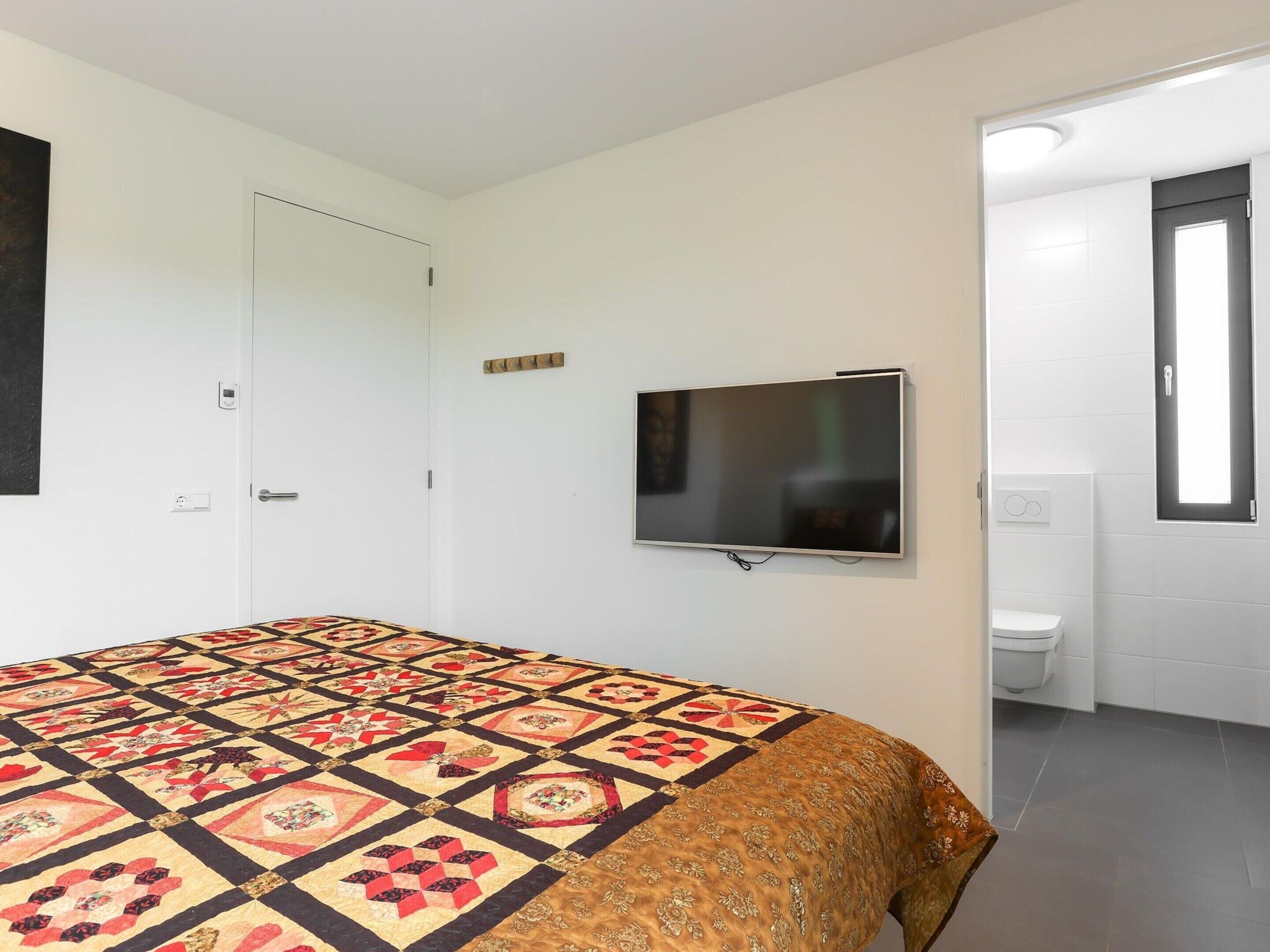A bedroom with a bed, a TV mounted on the wall, a door, and a bathroom visible through an open door.