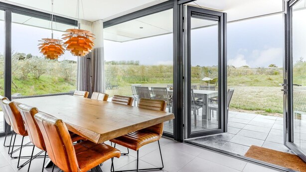 A modern dining area with a large wooden table and leather chairs, large windows, and a view of the outdoors. - Seaside Getaway With a Beautiful Garden (Kamperland)