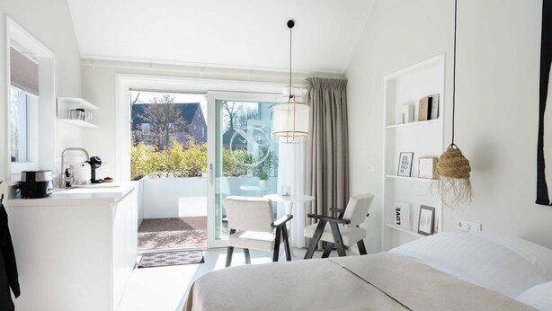 Una habitación de hotel moderna con una cama grande, un escritorio con cafetera, un balcón con vistas a zonas verdes y una lámpara colgante.
