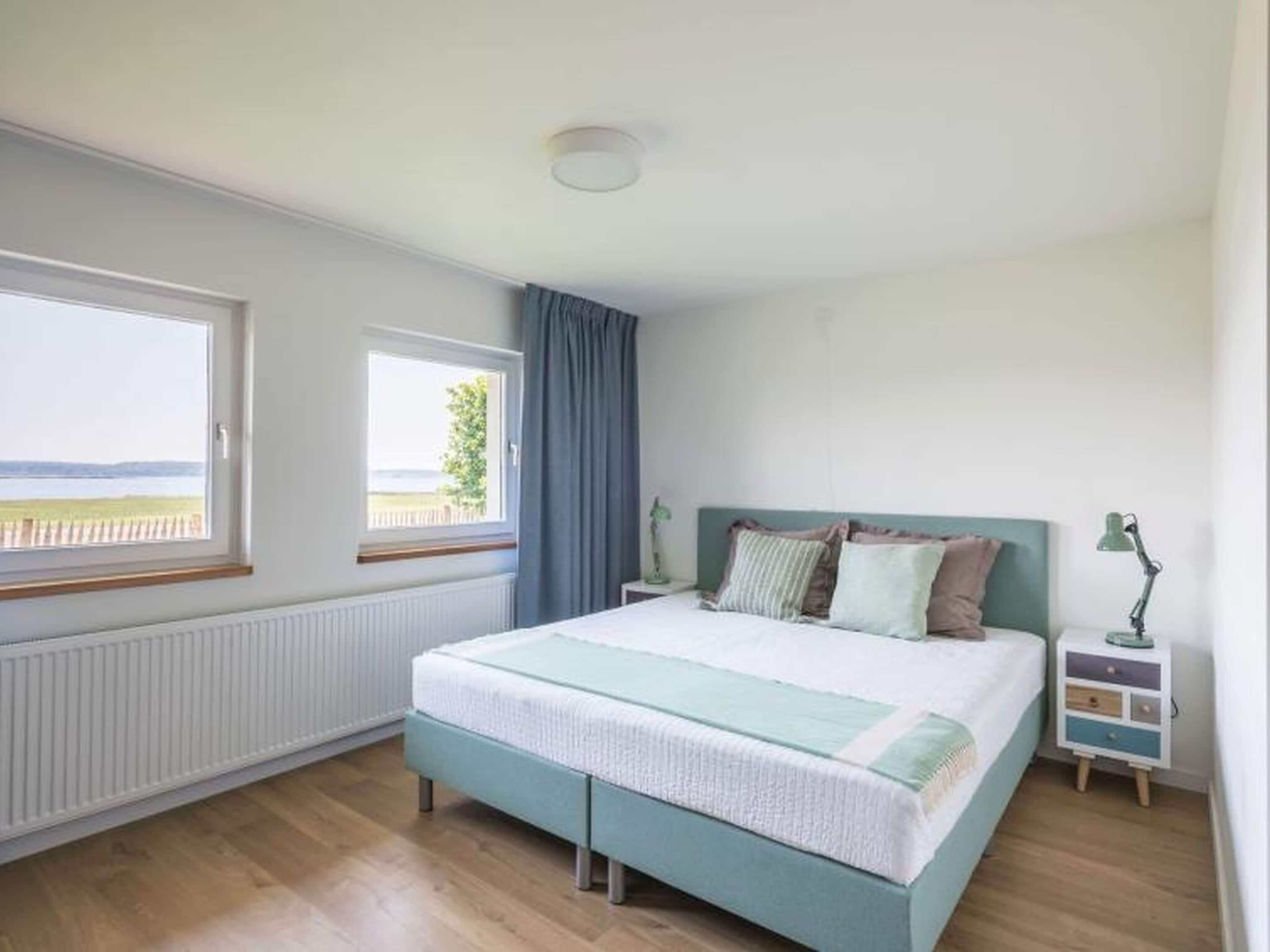 Een slaapkamer met een bed, twee ramen, een radiator, een nachtkastje en een lamp.