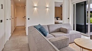 Een moderne woonkamer met een grijze bank, een ronde houten tafel en een grote schuifpui naar een buitenruimte.