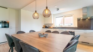 A modern kitchen with a wooden dining table, grey upholstered chairs, pendant lights, and a window with a view of a red-roofed building. - Beach Bliss Romantic Retreat (Scherpenisse)
