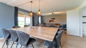 A modern dining area with a wooden table and grey upholstered chairs, a sofa, and a large window with curtains. - Beach Bliss Romantic Retreat (Scherpenisse)