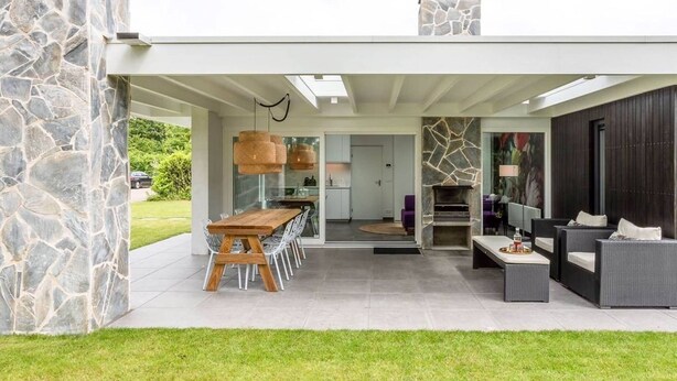 A modern outdoor patio with a stone wall, a wooden dining table, and a built-in fireplace. - Holiday Home Near Veerse Meer (Kamperland)