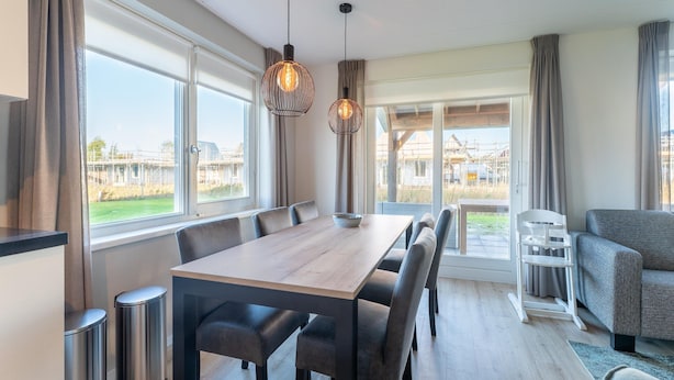 Un espace repas moderne avec une table et des chaises en bois, de grandes fenêtres avec des rideaux et une vue sur l’extérieur.
