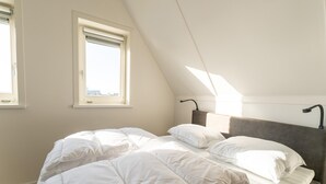 Une chambre avec un plafond en pente, deux fenêtres, un lit avec une literie blanche et une tête de lit dotée d’un luminaire.
