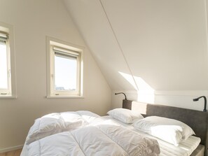 Un dormitorio con techo inclinado, dos ventanas, cama con ropa de cama blanca y cabecera con lámpara.