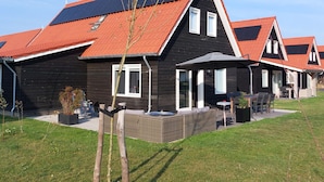 A row of modern houses with red roofs and solar panels, featuring outdoor seating areas and a well-maintained lawn.