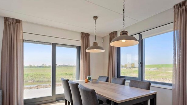 A modern dining room with a large table and chairs, large windows with curtains, and pendant lighting.