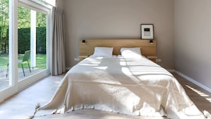 Une chambre avec une grande fenêtre, un lit recouvert de draps blancs, une tête de lit en bois et un tableau encadré accroché au mur.
