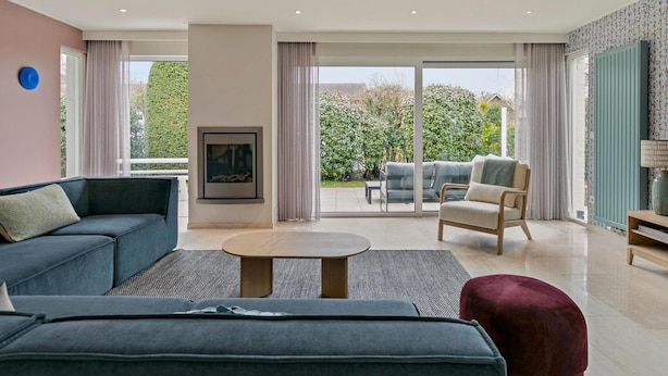 Un salon moderne comprenant un canapé, un fauteuil et un pouf, une table basse, et donnant sur un jardin grâce à de grandes portes-fenêtres coulissantes.