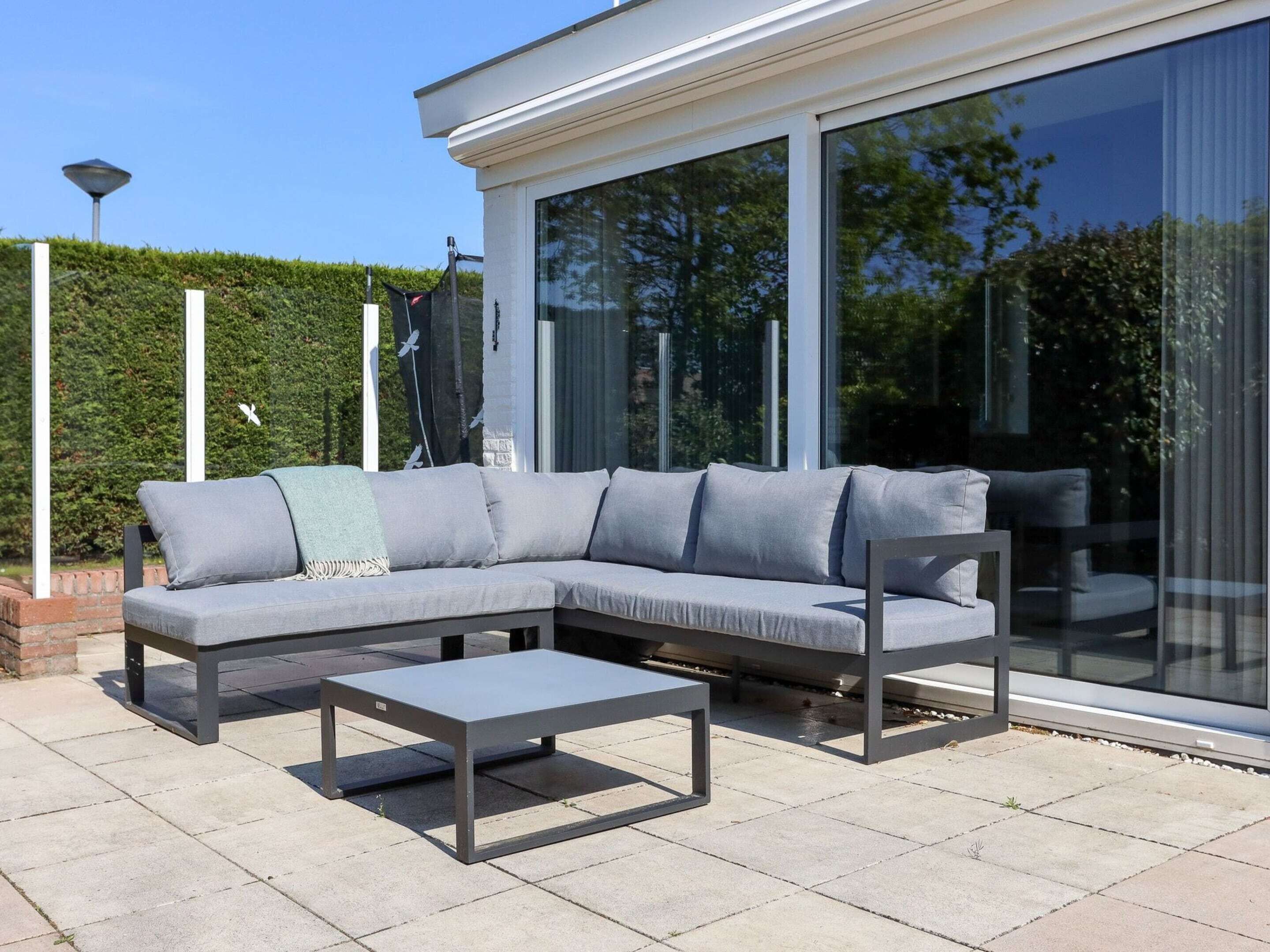 A modern outdoor seating area with a grey sectional sofa and a metal coffee table, set on a paved patio with a large glass sliding door in the background.