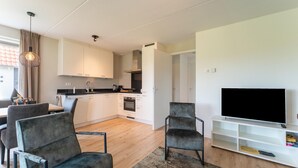 Una sala de estar moderna con televisor, dos sillones, una mesita de centro y una cocina al fondo.