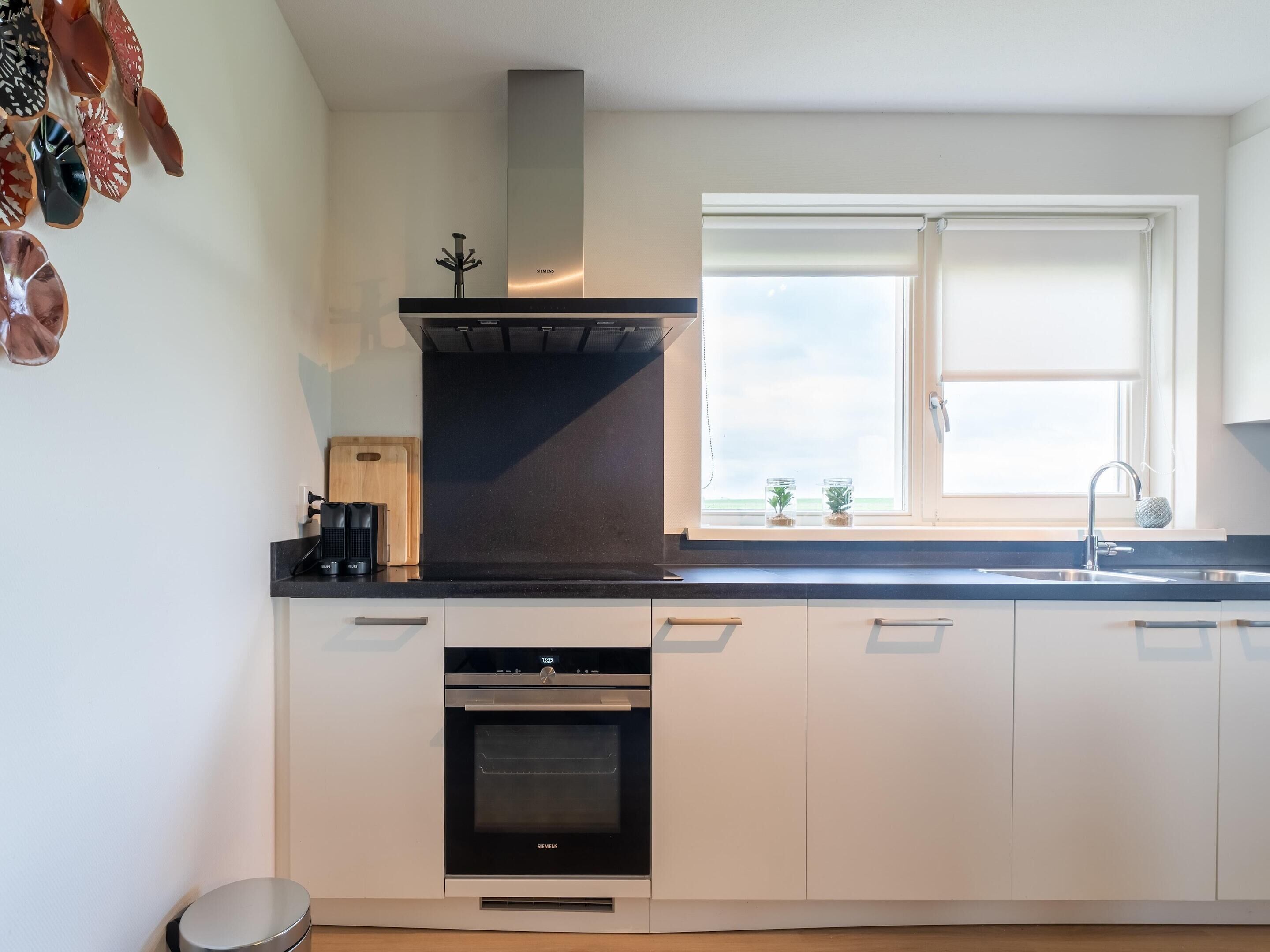 A modern kitchen with white cabinets, a black oven, a stainless steel range hood, a window with blinds, and a sink with a faucet.
