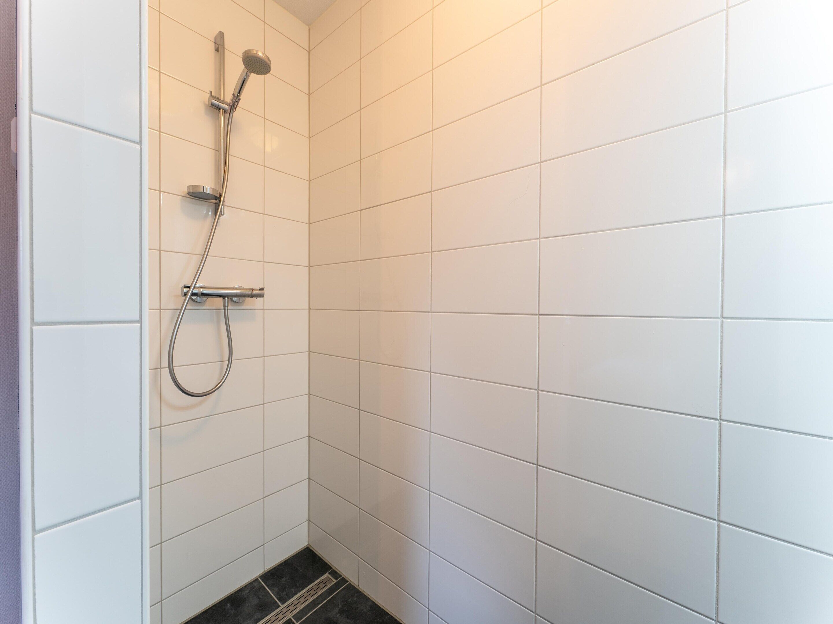 Un baño con paredes de azulejos blancos, una ducha con cabezal de ducha manual y un desagüe en el suelo.